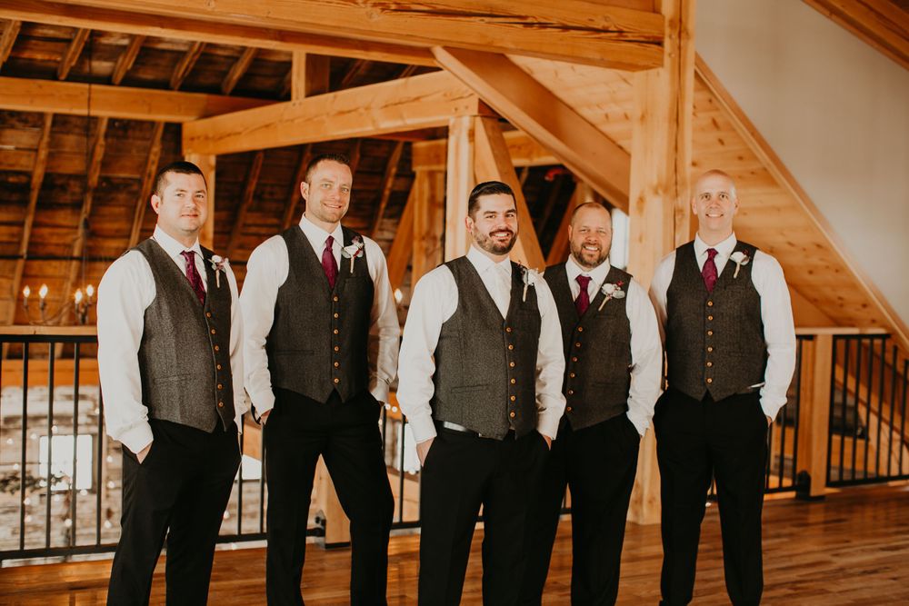 Five groomsmen in gray vests and burgundy neckties pose inside a warmly lit rustic barn venue.