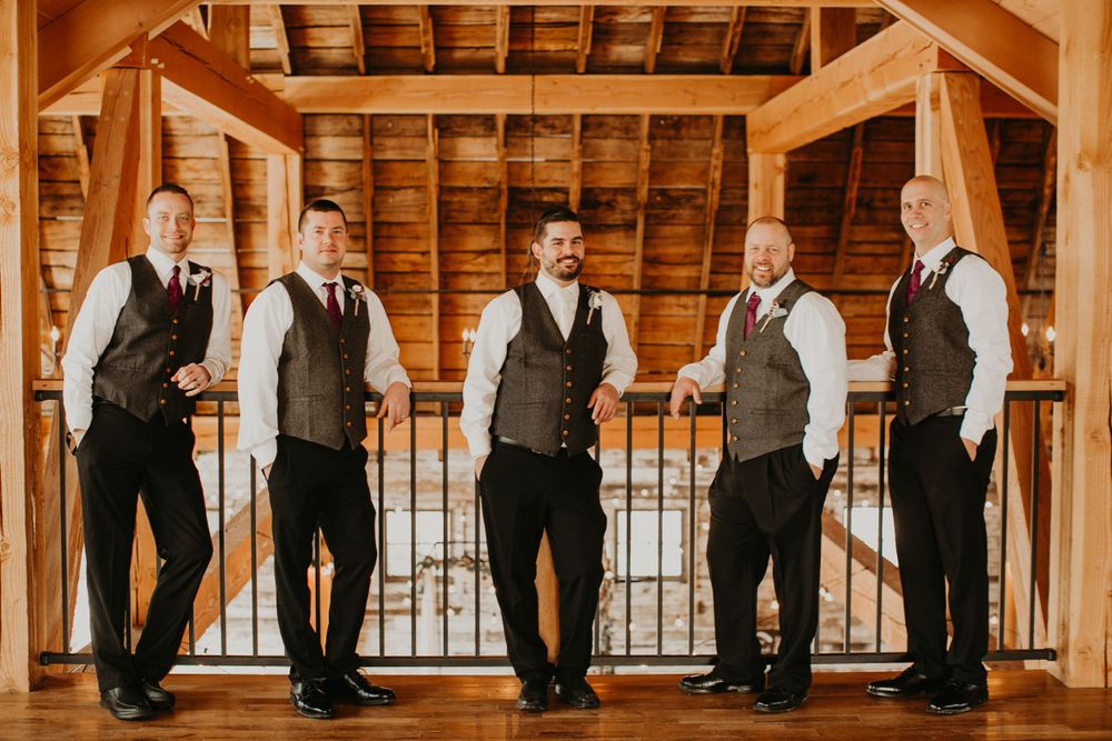 Groomsmen in matching gray vests lean casually on a balcony railing inside a rustic barn venue.