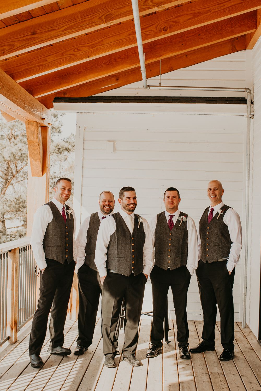 Five groomsmen in gray vests and dark pants pose on a wooden porch at a rustic wedding venue.