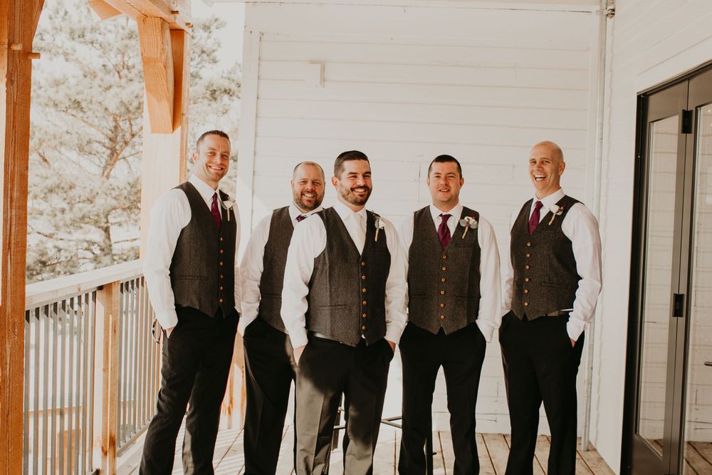 Five groomsmen in matching gray vests smile together on a wooden porch at a rustic wedding.