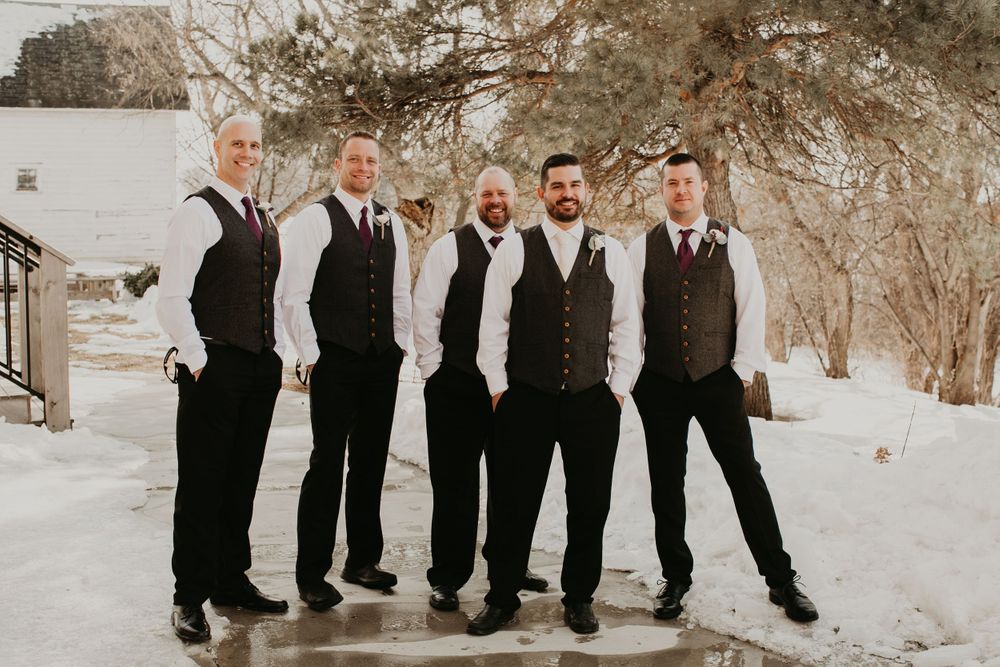 Six groomsmen in gray vests and dark pants pose together outdoors in a snowy winter setting.