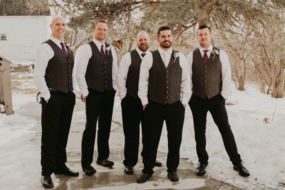 Five groomsmen in matching gray vests and black pants pose together outdoors in a snowy winter wedding setting.