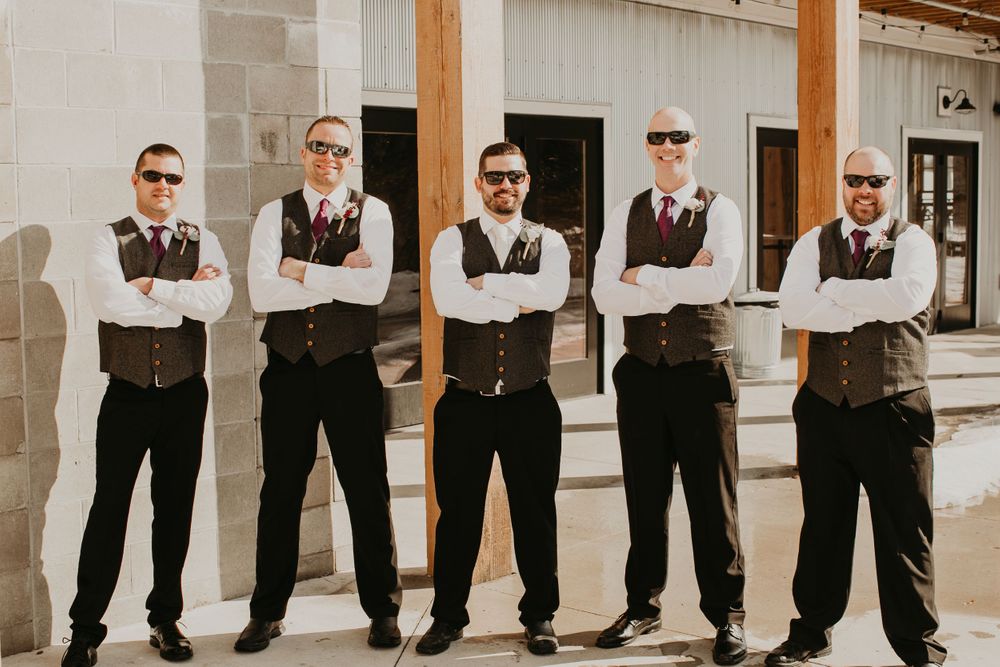 Five groomsmen wearing sunglasses and gray vests cross their arms confidently outside a stone wedding venue.