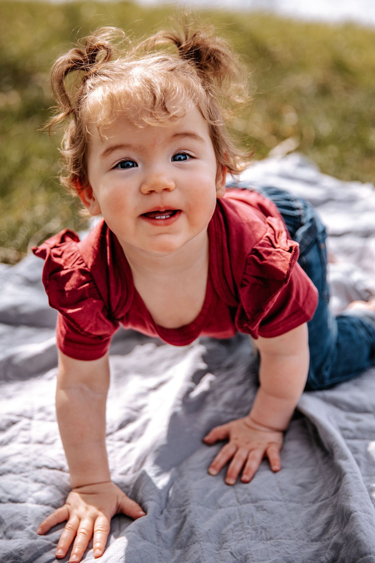 A smiling toddler in a red shirt crawls on a blanket outdoors.