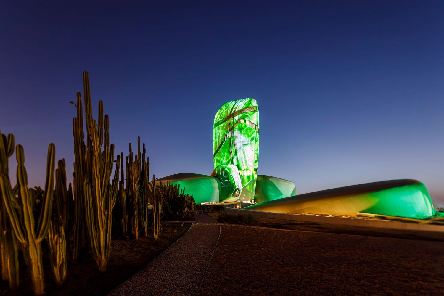 Illuminated green glass sculpture alongside desert cacti at dusk.