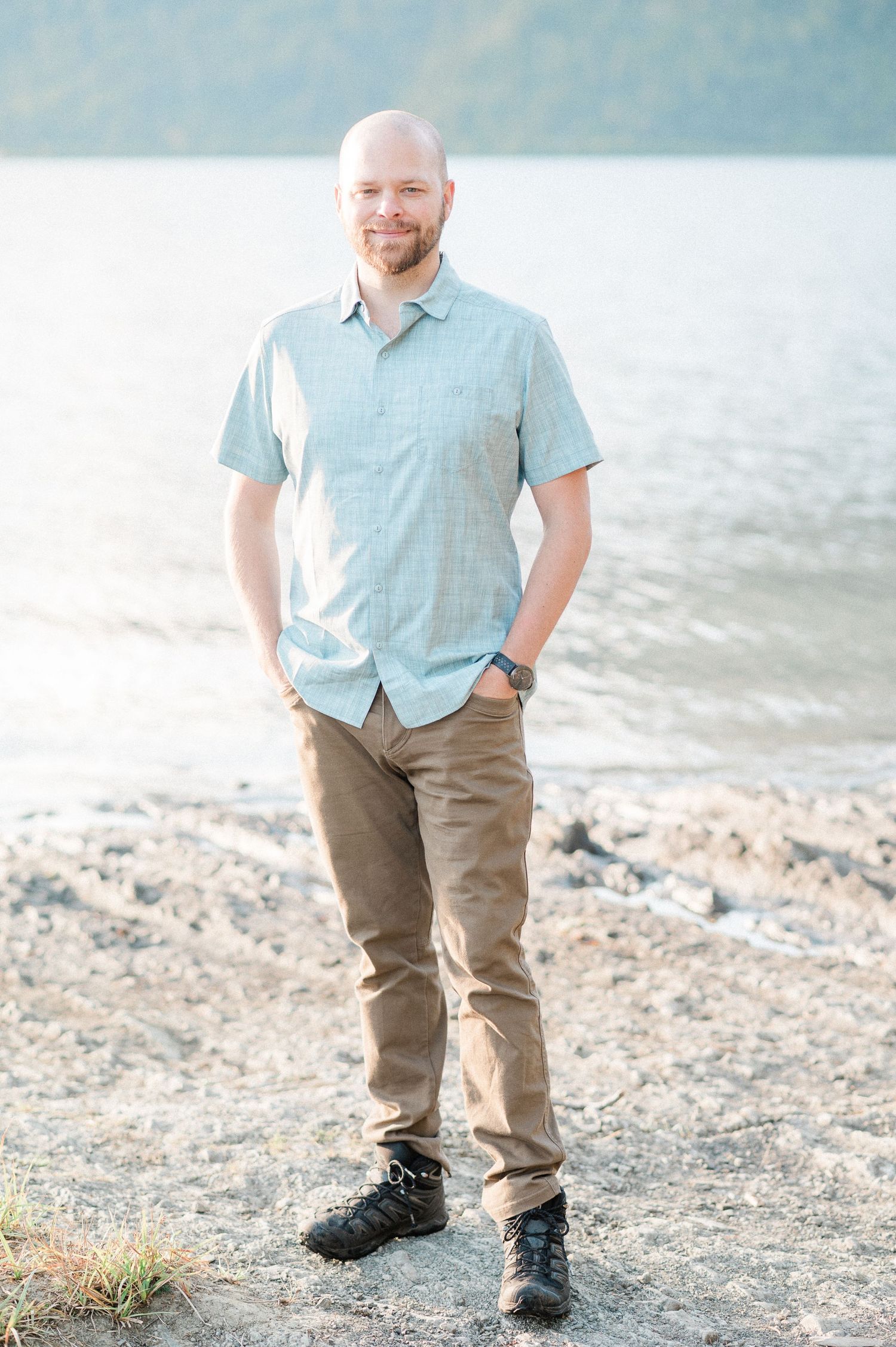 A person in a light blue button-down shirt and khaki pants stands on a rocky beach shoreline at sunrise