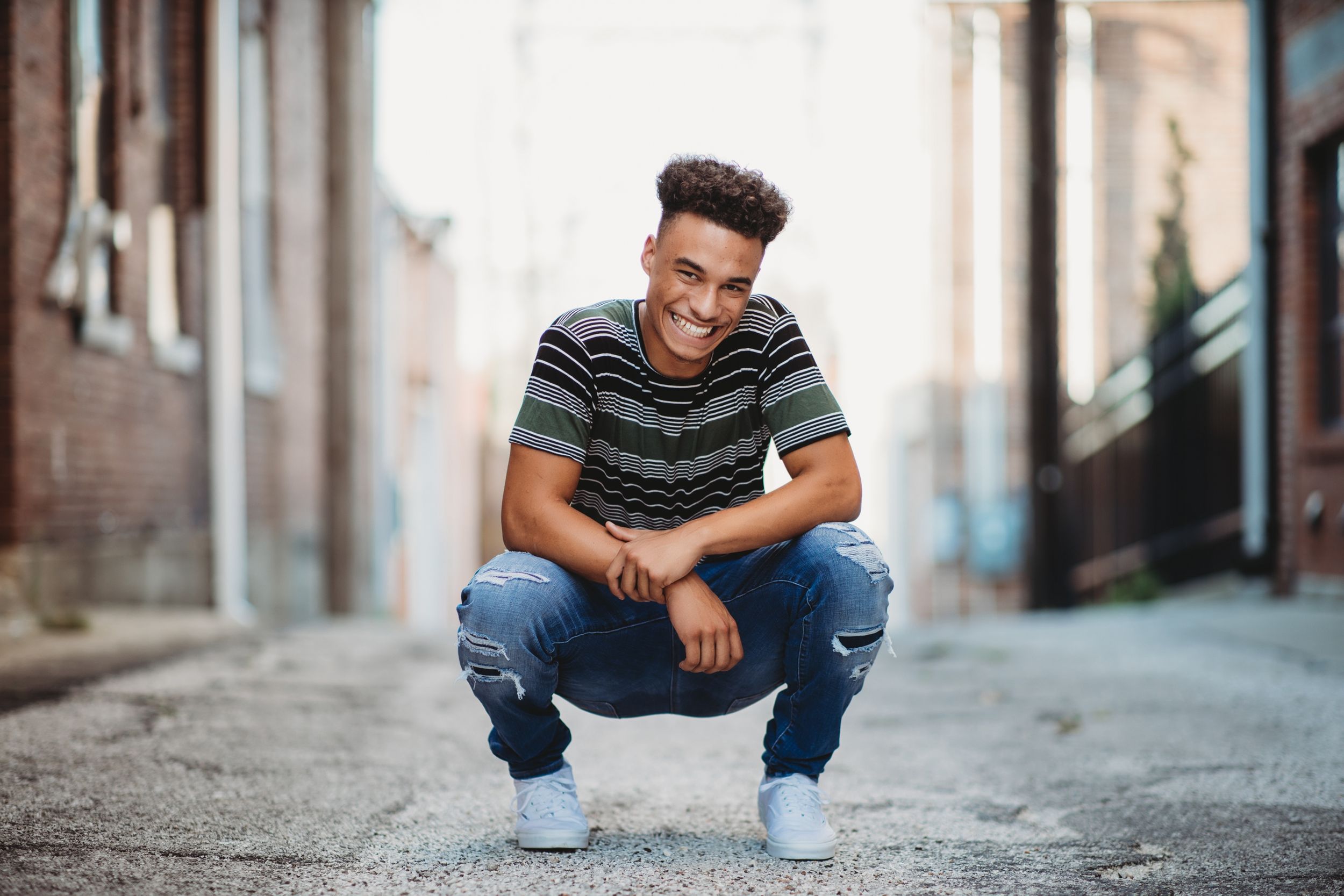 Casual portrait of a young man in ripped jeans and striped shirt crouching in an urban alley.