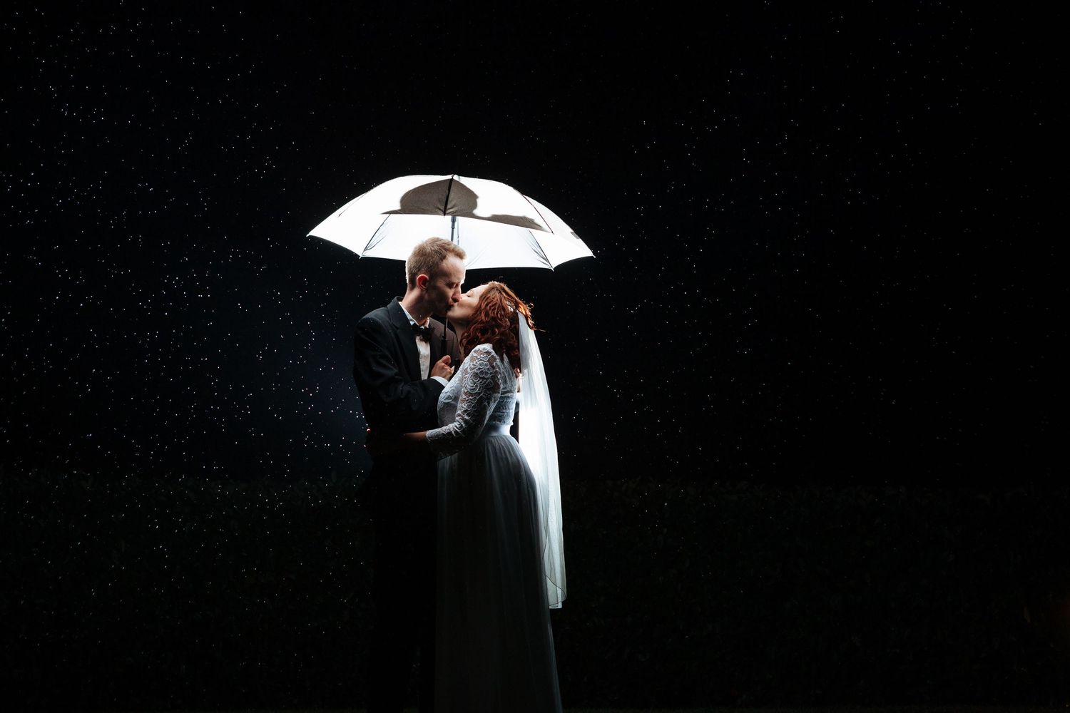 A bride and groom stand under a light umbrella in the rain and dark.