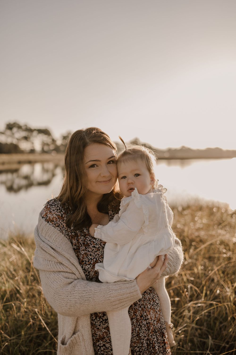 Golden hour portrait of parent and baby by a peaceful lake with warm sunset lighting.