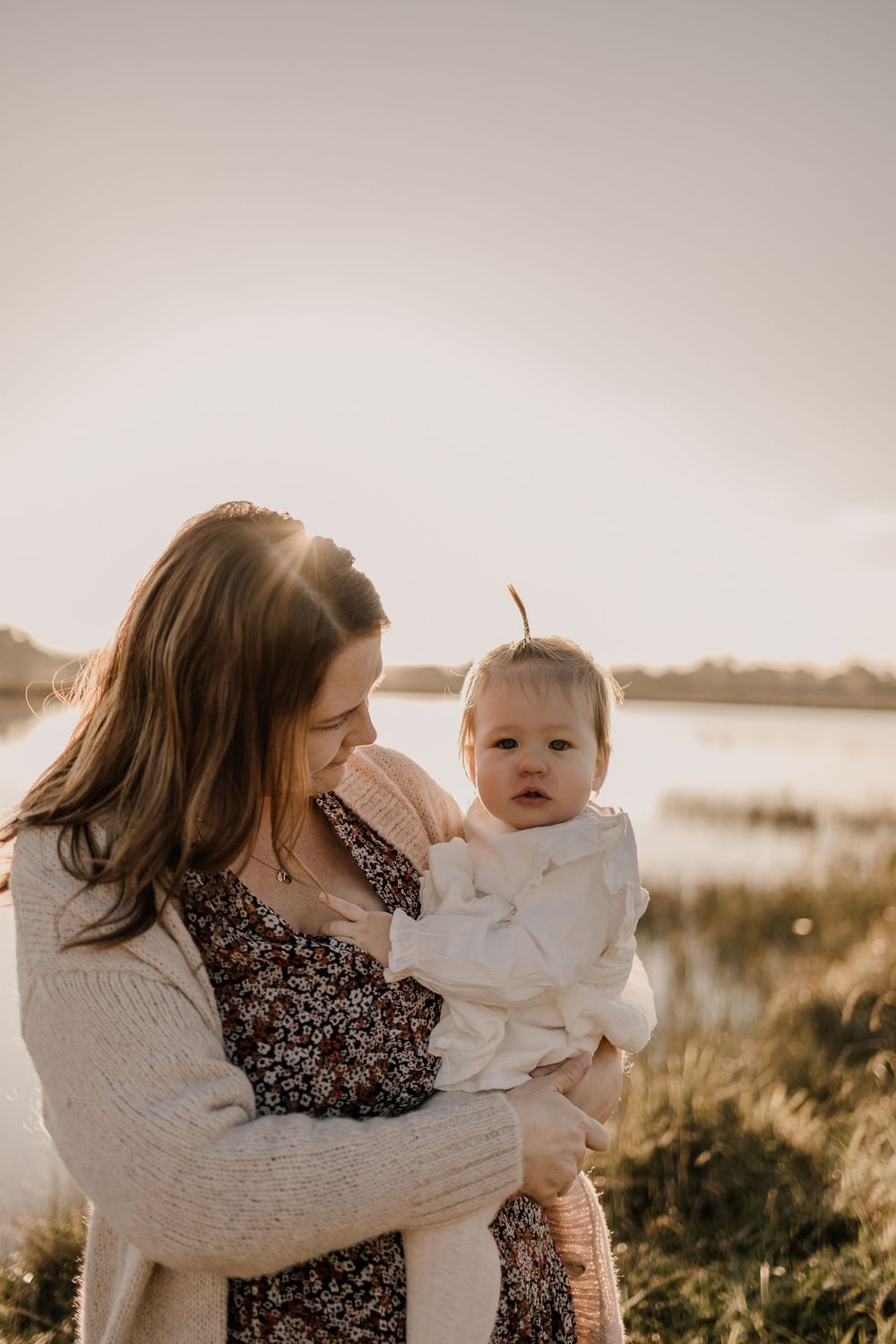 Parent holding baby in white dress against sunset backdrop near waterfront.