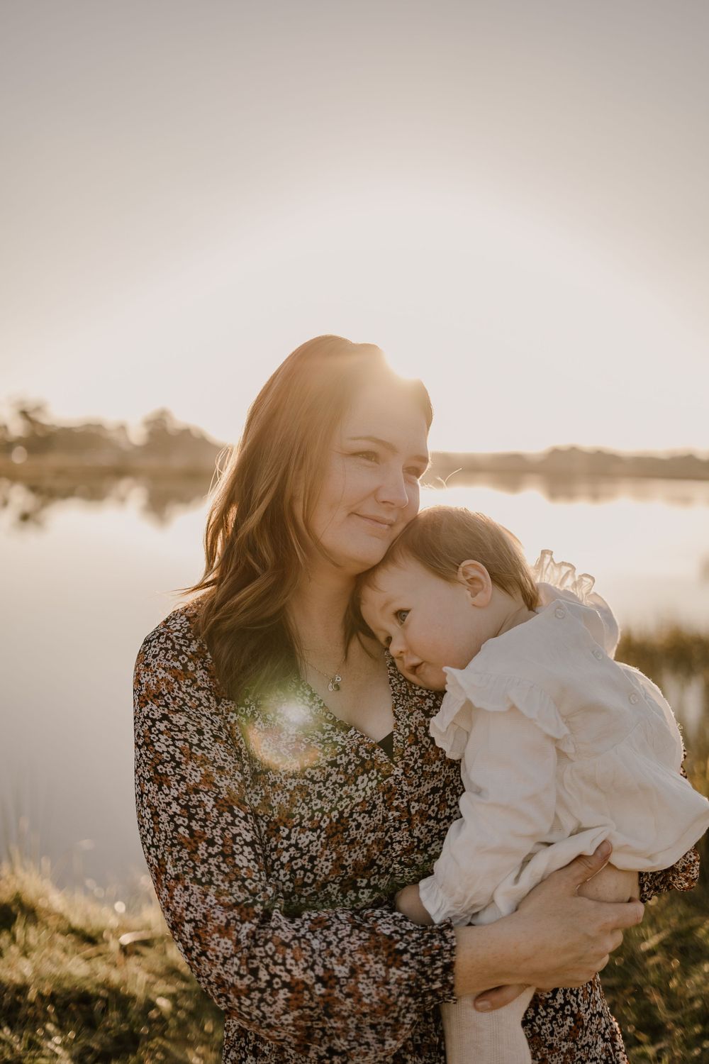 Sun-drenched portrait of parent and baby embracing by lakeside during golden hour.
