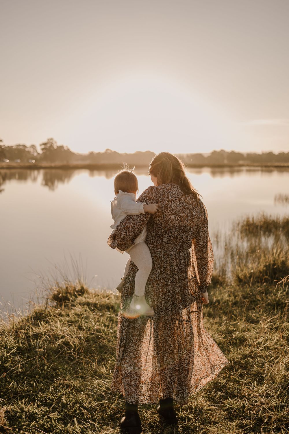 Silhouette of parent holding child while watching sunset by tranquil lake.