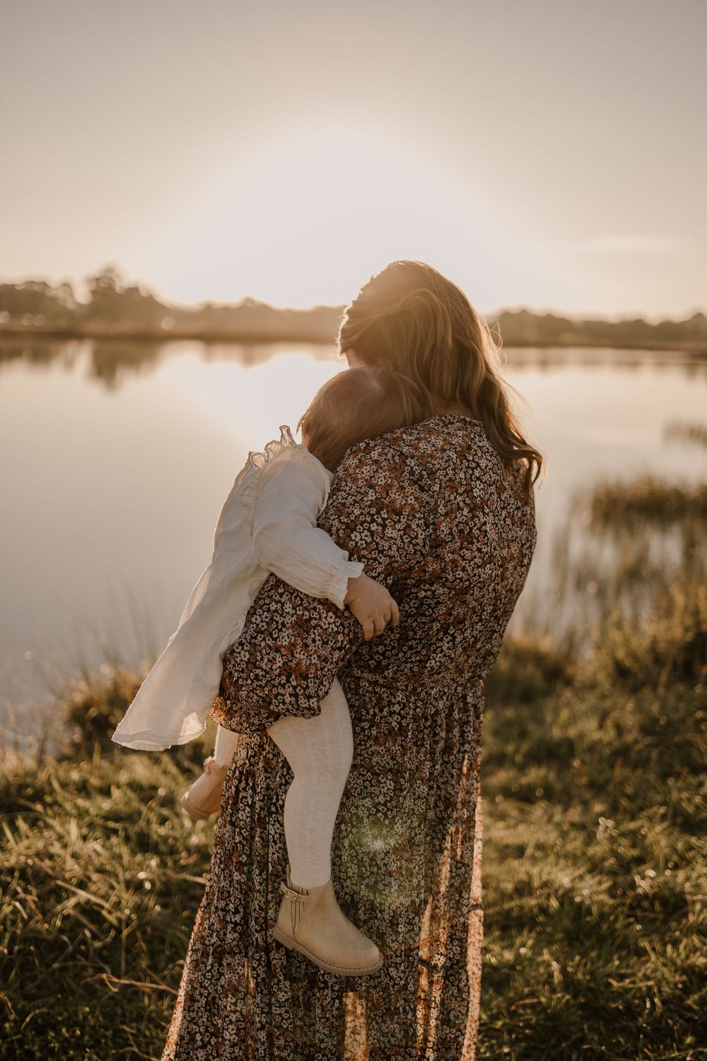 Two figures embrace by a tranquil lake at sunset, casting long shadows on the grassy shore.