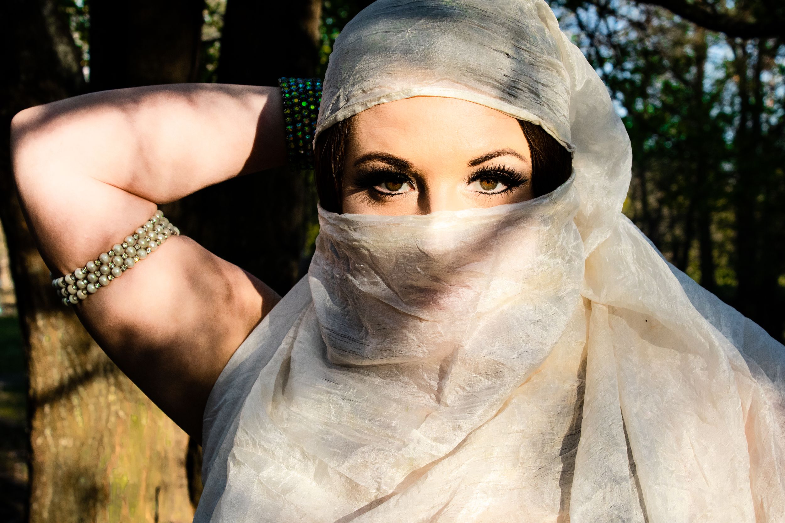 A bride in a white wedding veil poses outdoors in a natural setting with sunlit trees in the background.