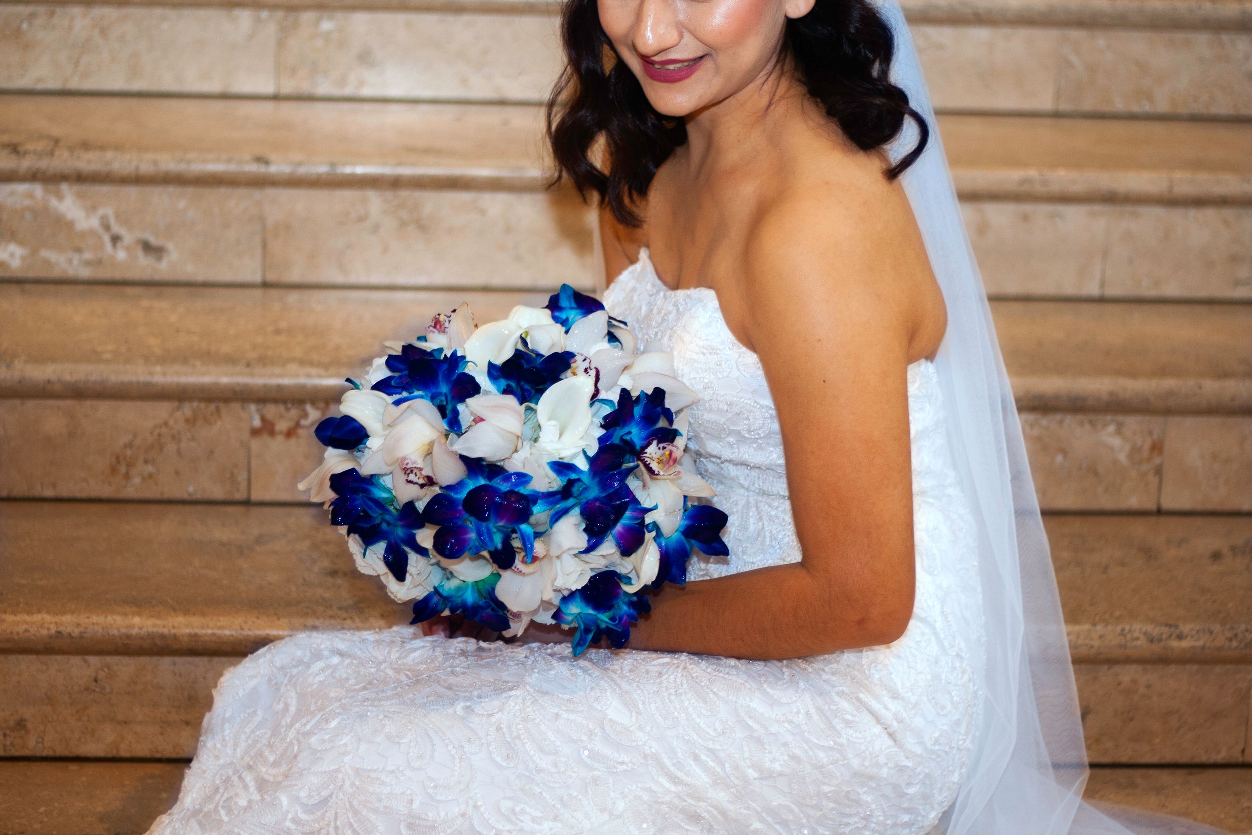 A bride in a white wedding dress holds a vibrant blue and white orchid bouquet against a rustic wooden wall backdrop.