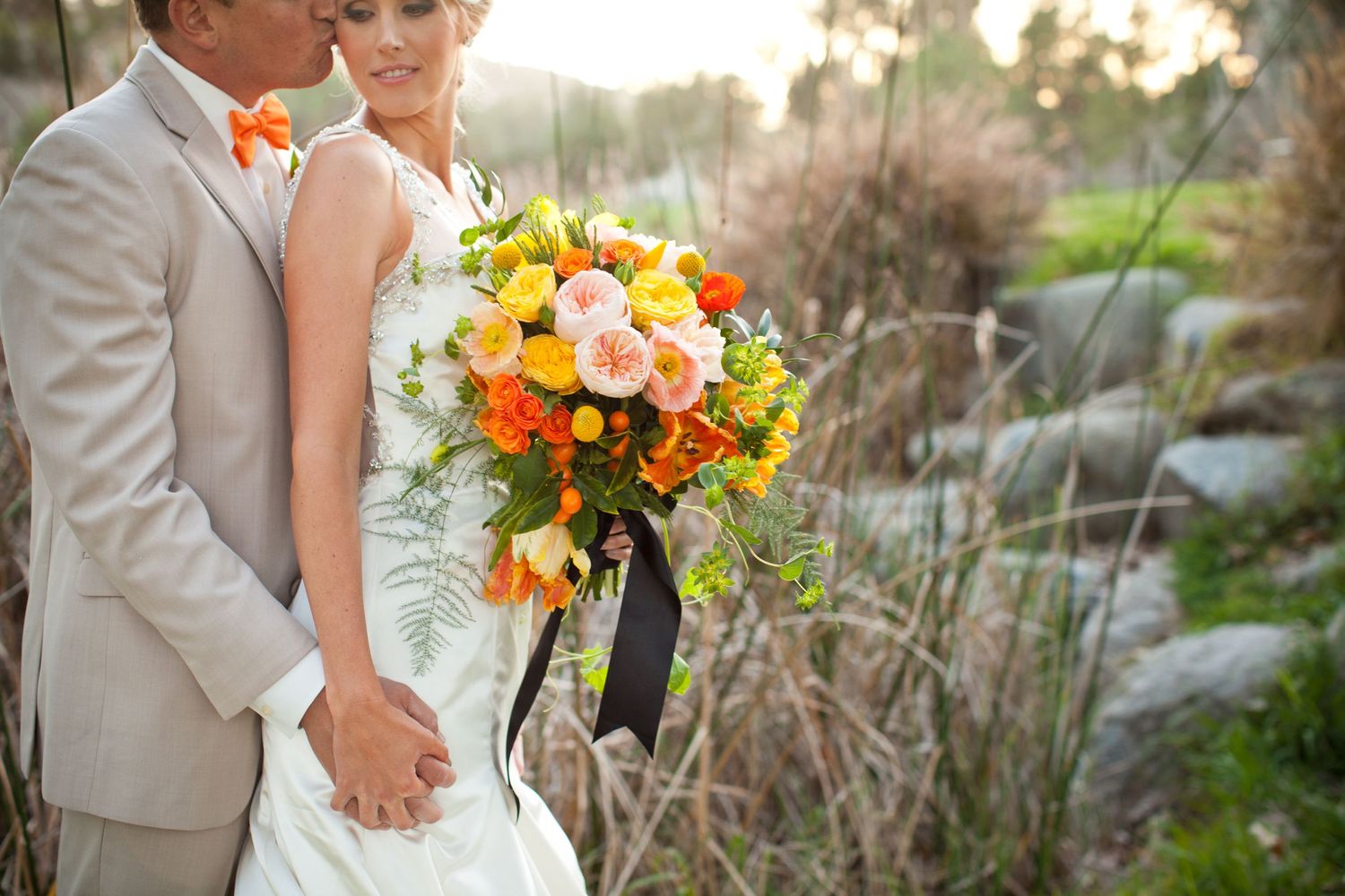 groom kissing bride and holding a orange and yellow bouquet on a golf course