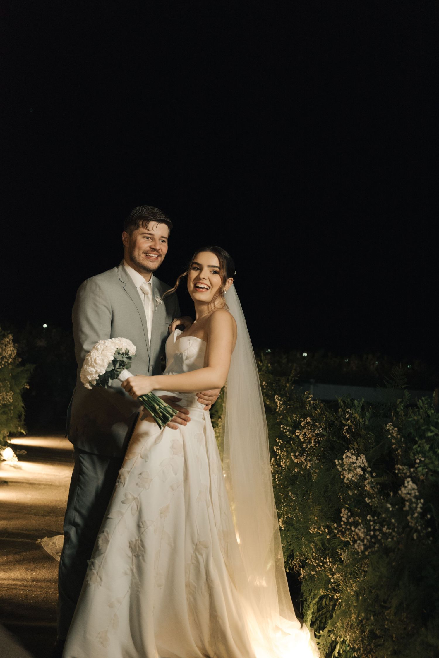 Evening wedding portrait with couple holding bouquet on illuminated garden path.