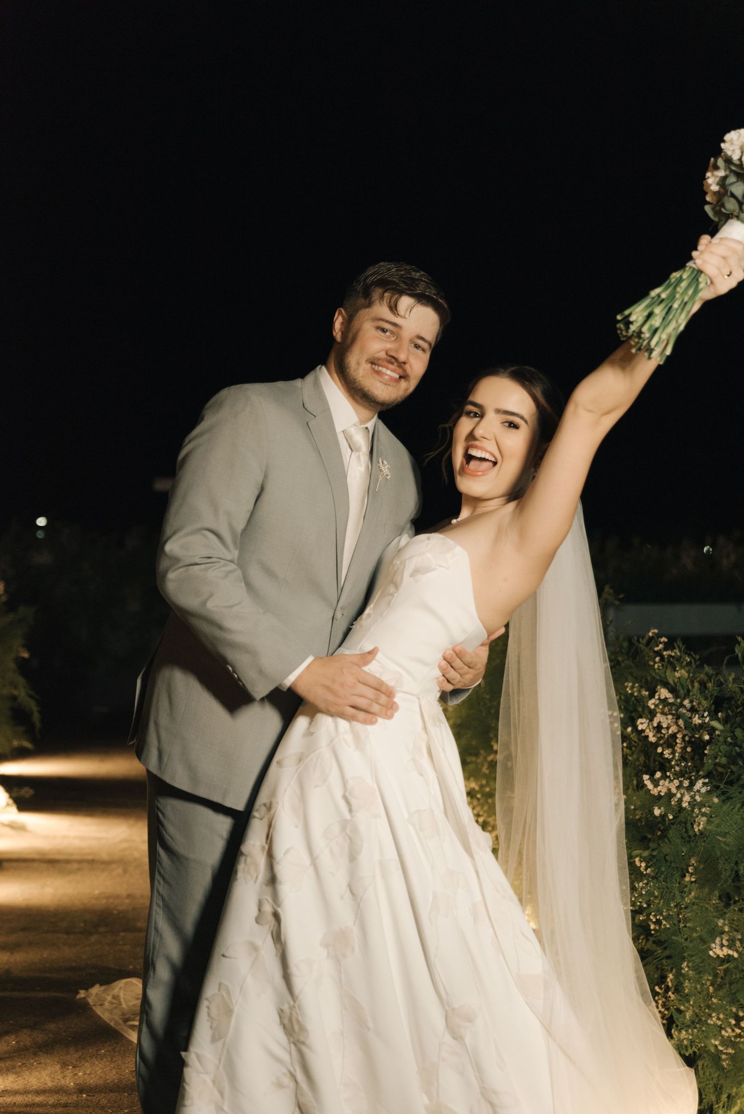 Joyful wedding couple celebrating outdoors with raised bouquet at night.