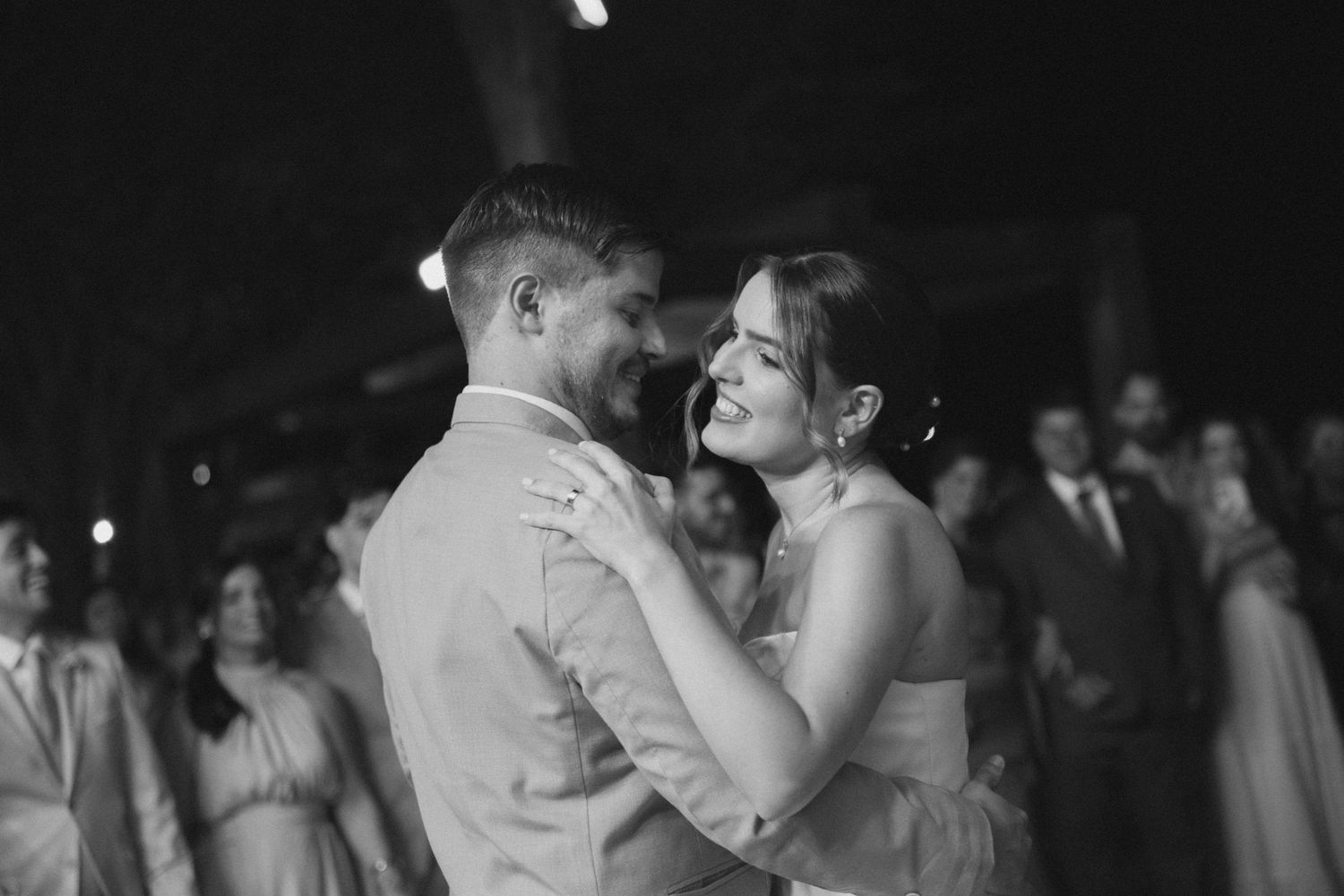 Intimate black and white moment of newlyweds dancing closely together surrounded by wedding guests.