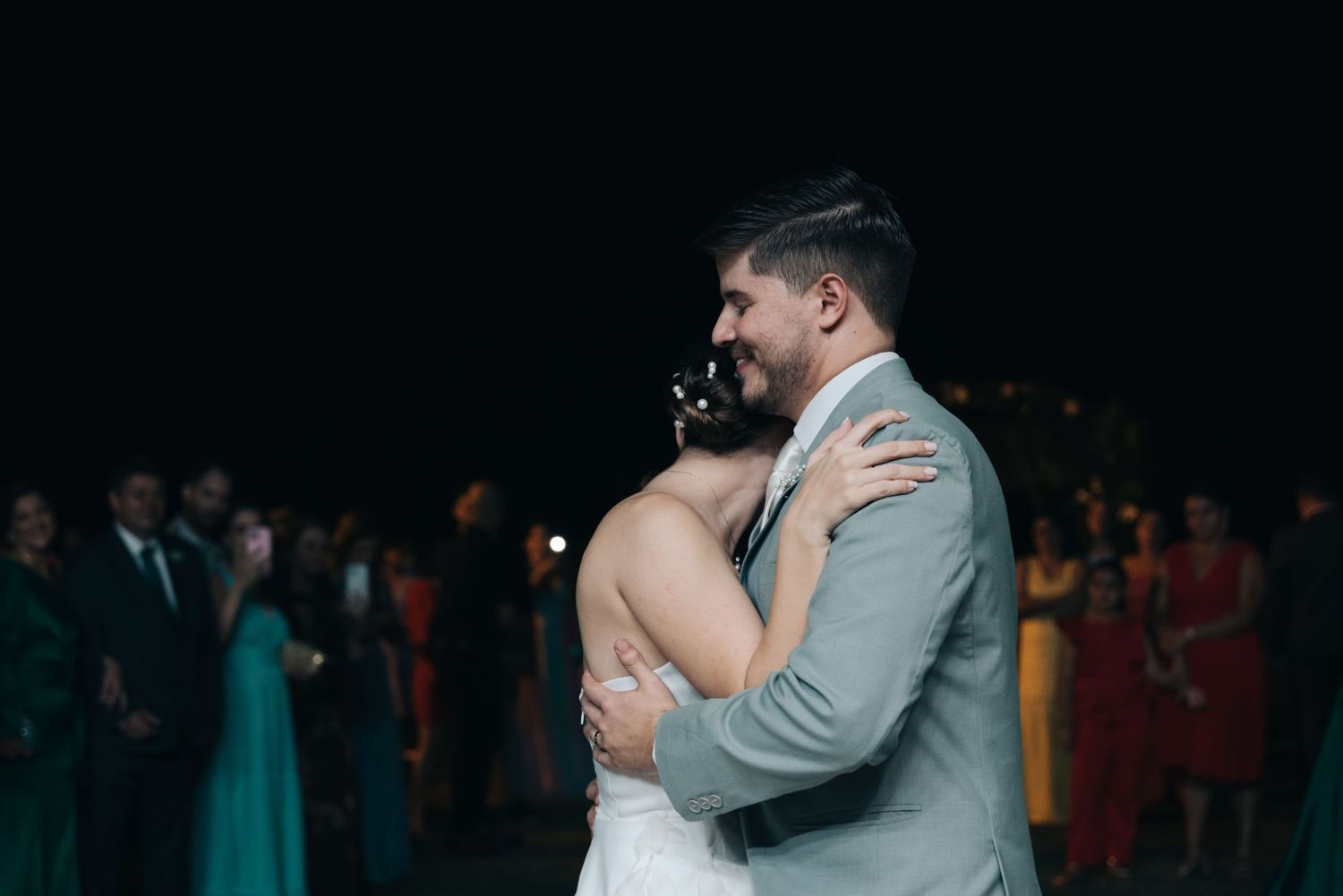 Couple embraces during wedding dance with guests watching in colorful formal attire.