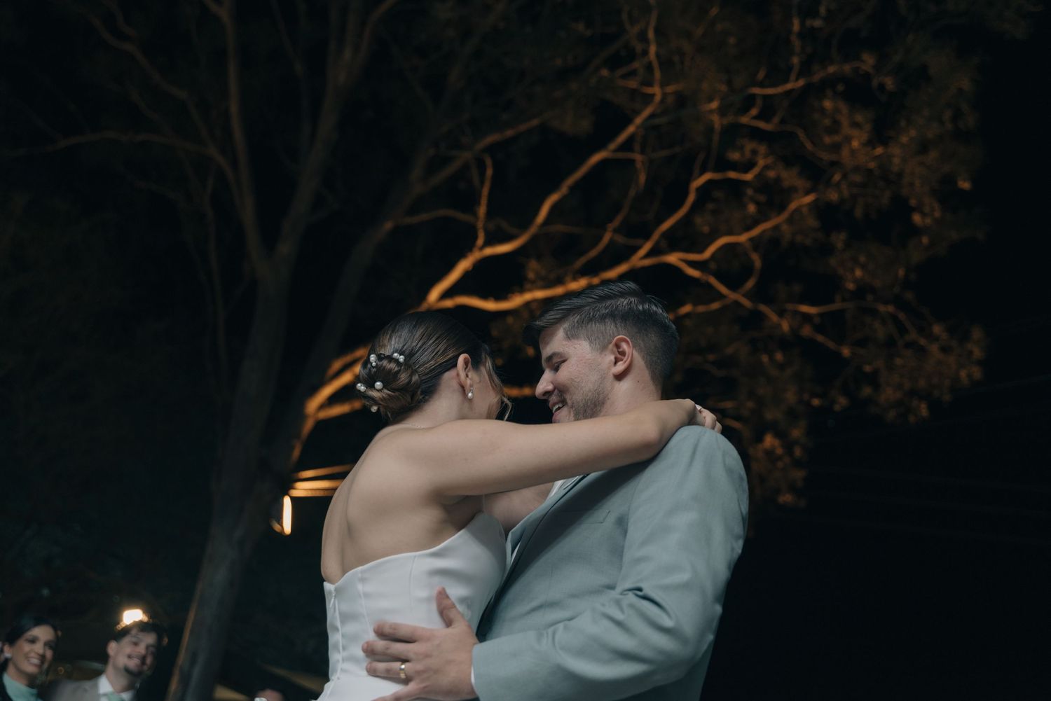 Romantic nighttime wedding dance with couple embracing under illuminated tree branches.