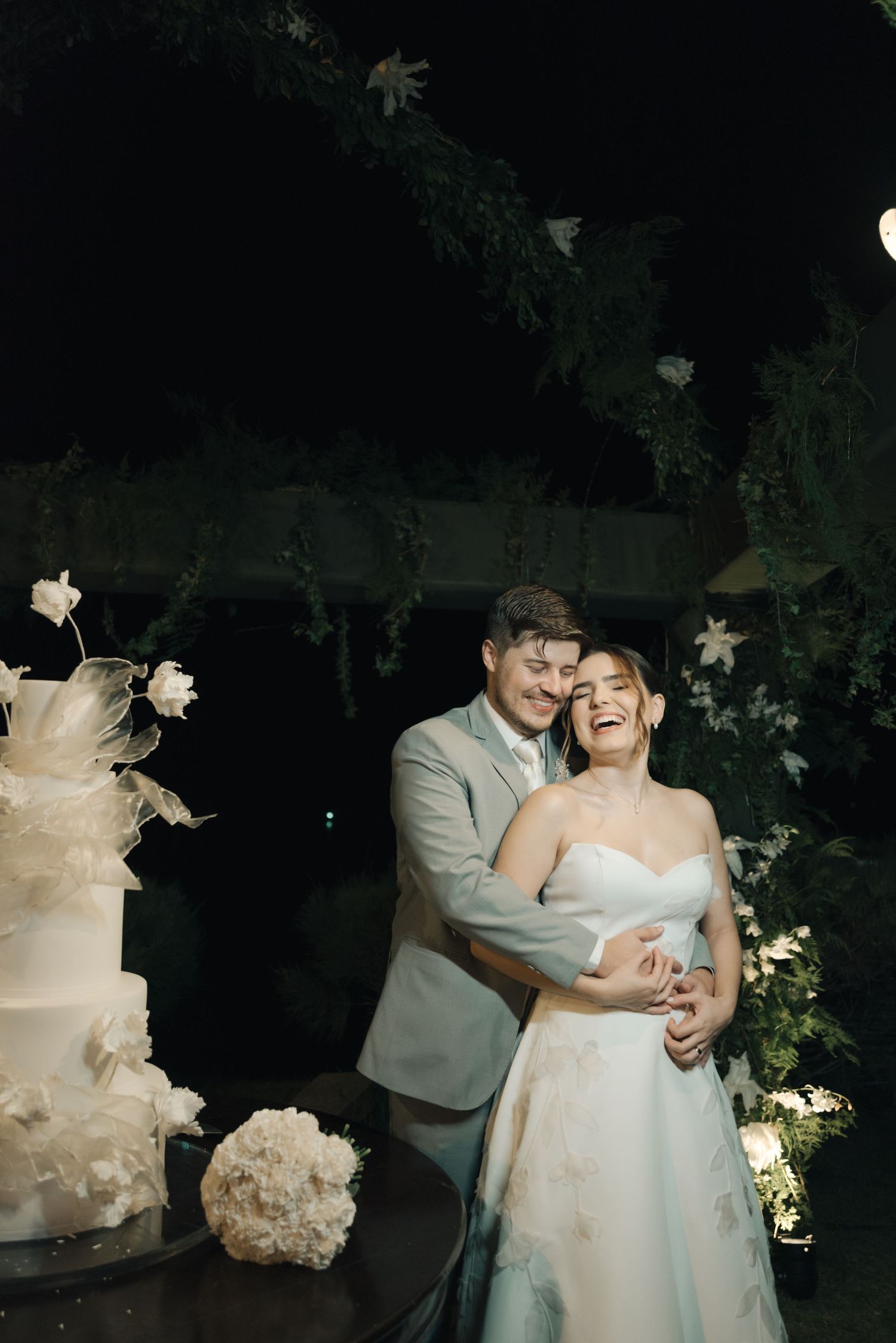 Joyful wedding moment captured next to elegant white wedding cake with floral decorations.