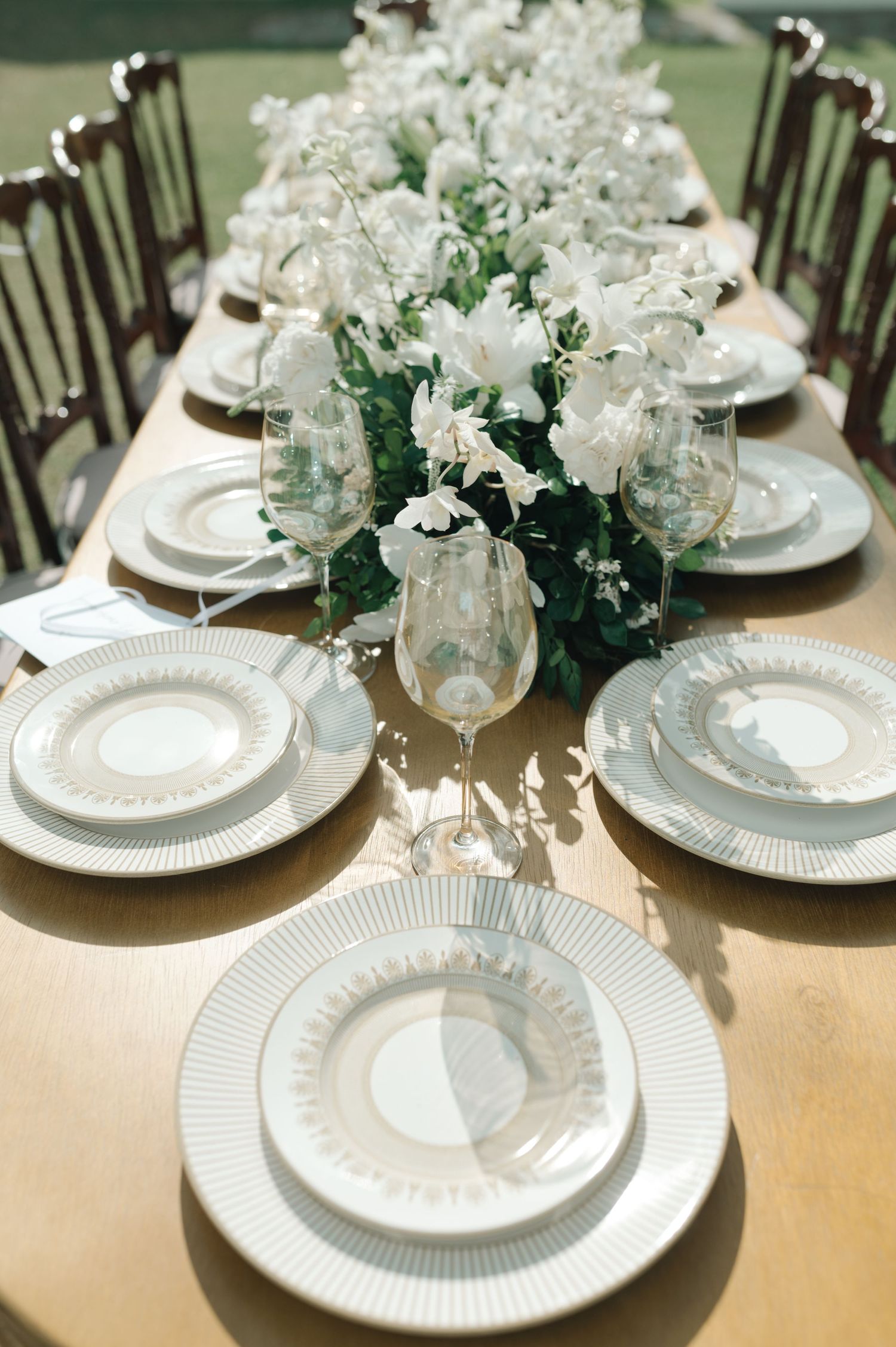 Elegant wedding table setting with white plates, floral centerpieces and dark wooden chairs.