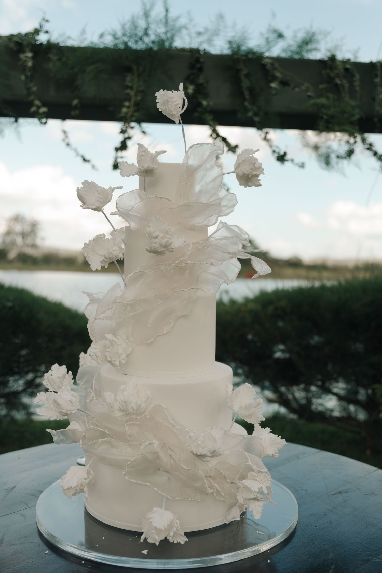 Elegant white wedding cake decorated with delicate sugar flowers against outdoor backdrop.