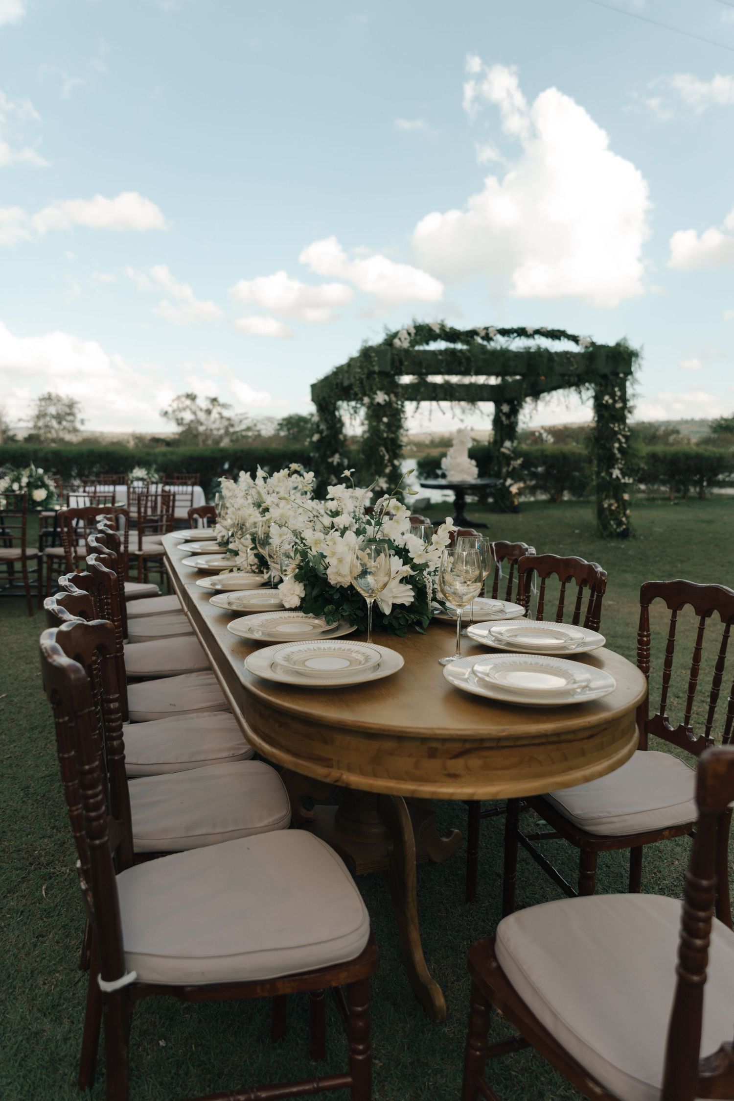 Long wooden dining table with place settings under green-covered pergola at outdoor wedding.