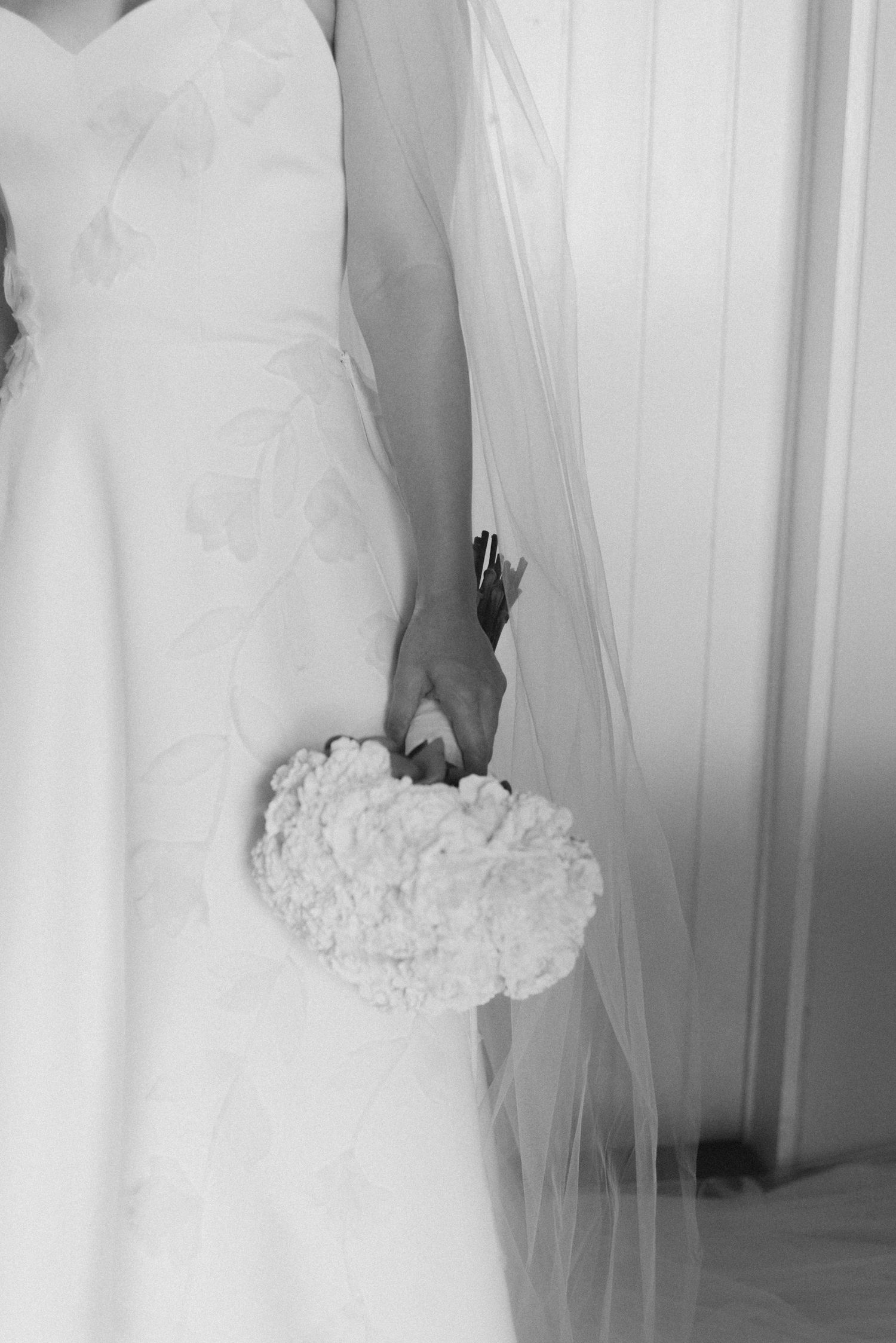 Black and white detail shot of bride holding white flower bouquet against white wall.