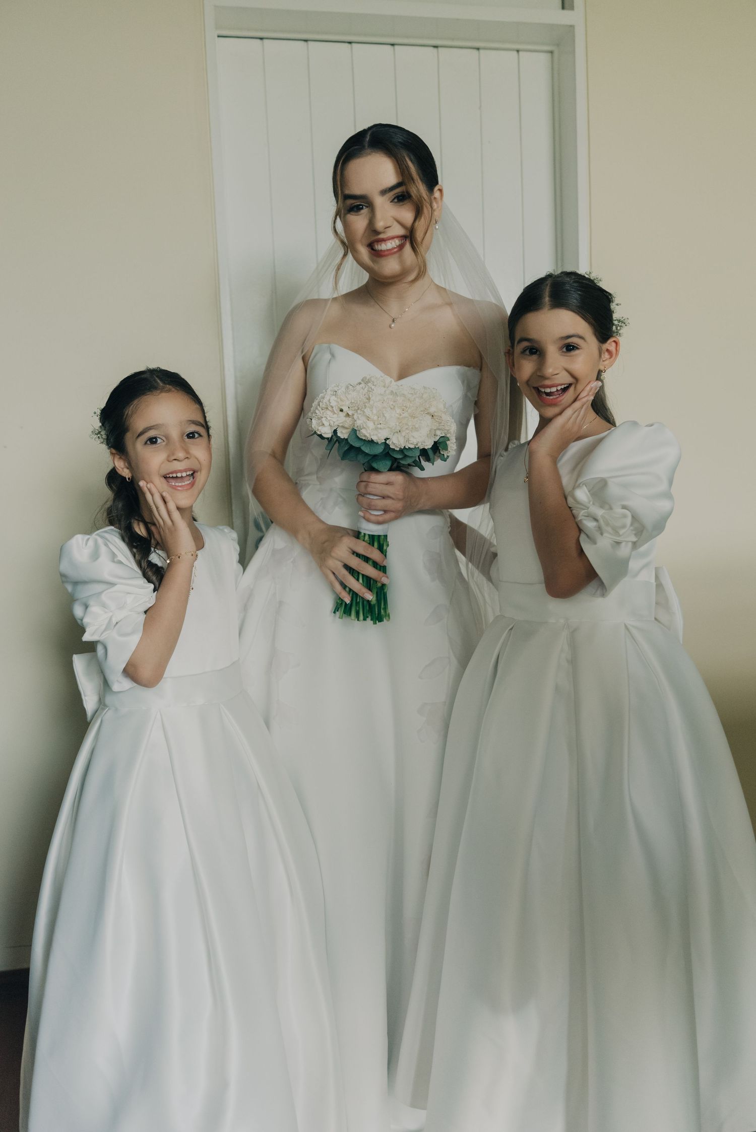 Three figures in elegant white gowns pose together for a wedding photo, with the bride holding a blue bouquet.