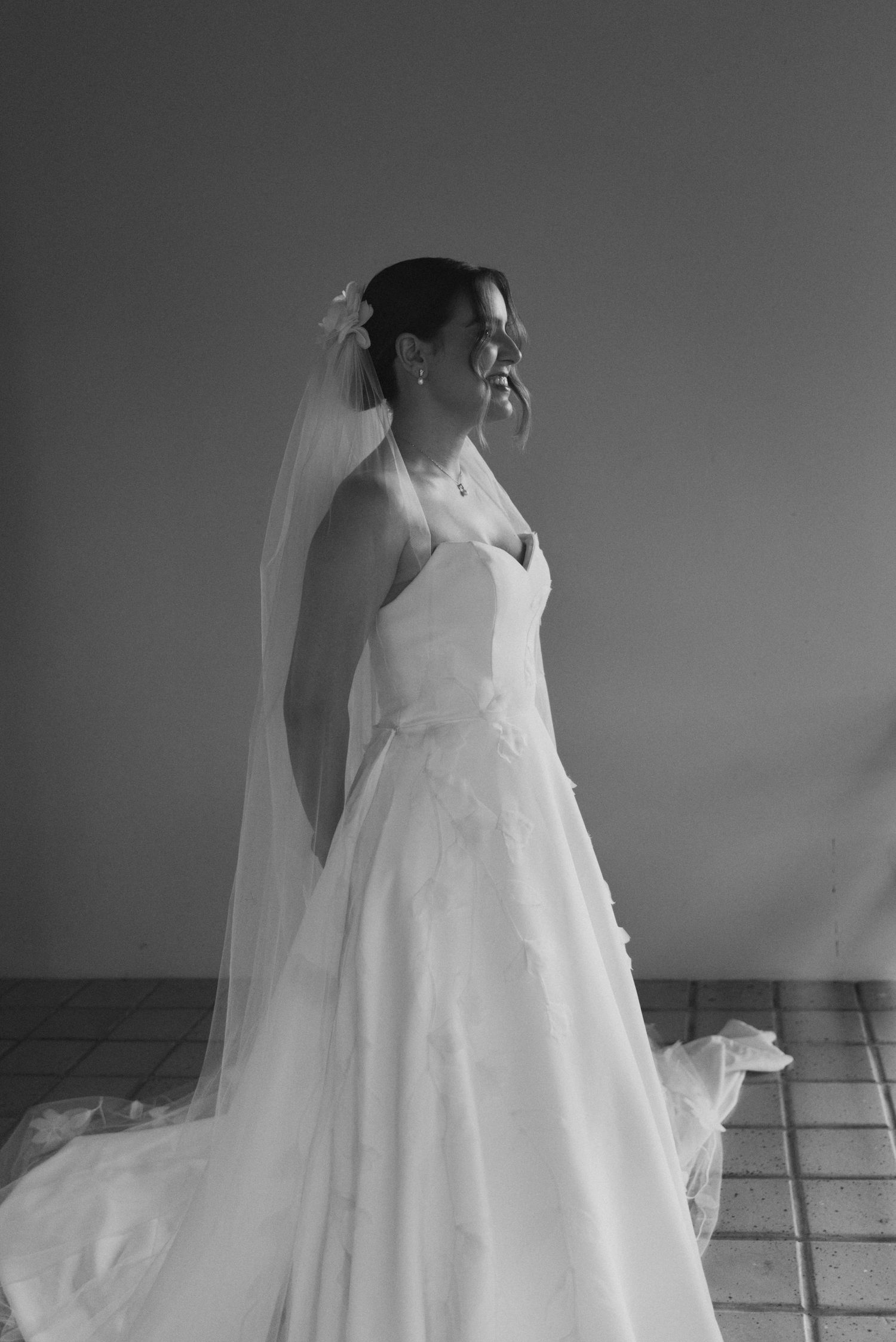 A side profile of a bride in a classic white wedding gown with a flowing veil against a neutral background.