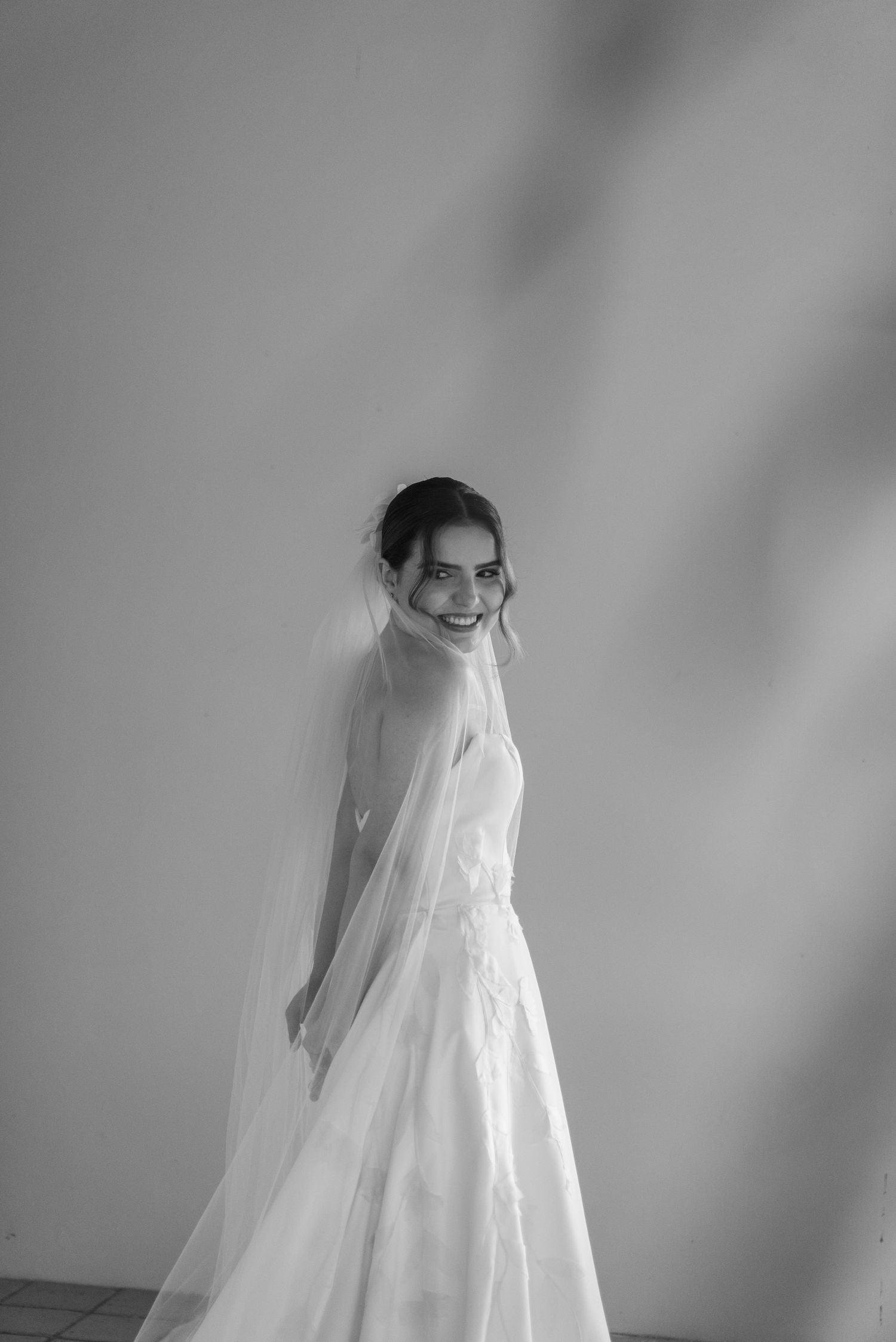 A bride in a classic white wedding dress and veil smiles while posing against a light background.
