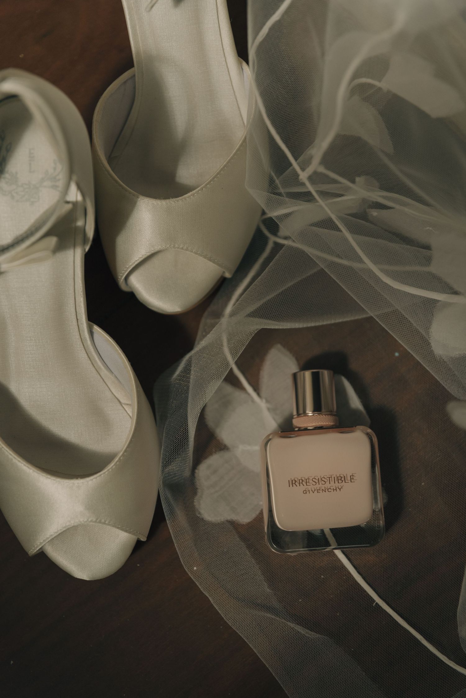 Wedding accessories including white peep-toe heels and perfume bottle arranged with delicate veil.