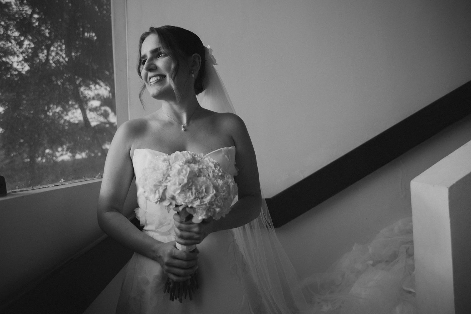 Black and white photo of bride holding white bouquet while standing near window in wedding dress.