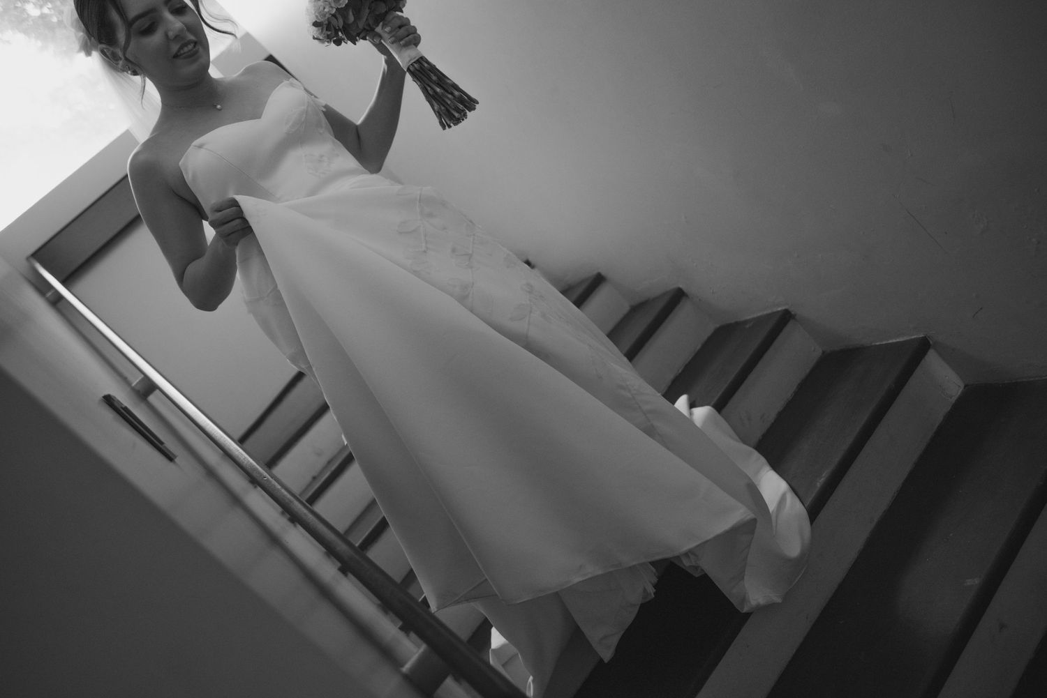 Artistic black and white shot of bride descending staircase with flowing dress and bouquet.