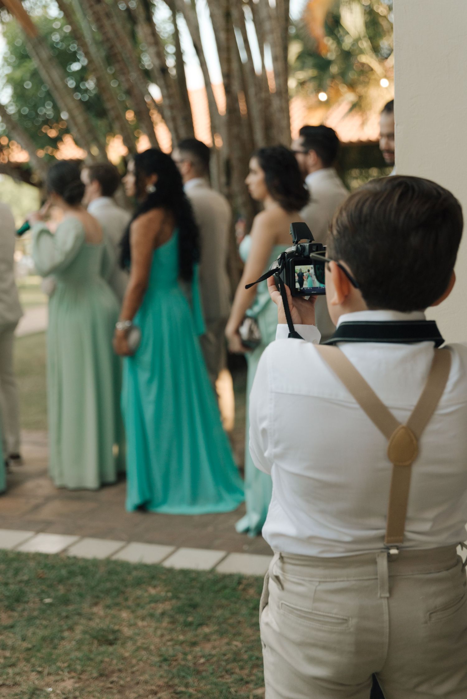 Young photographer in suspenders capturing bridesmaids in turquoise dresses at outdoor ceremony.