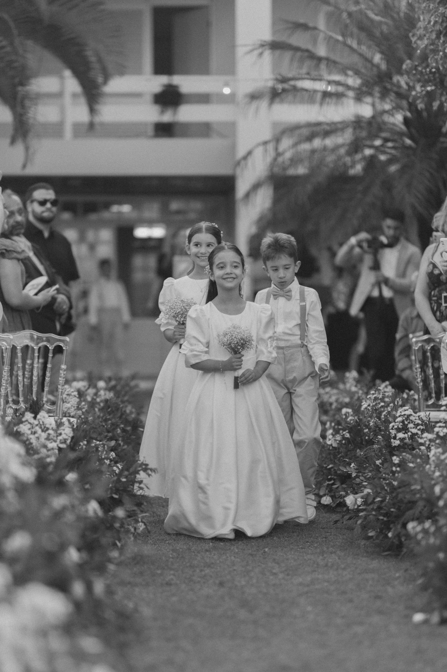 Young attendants walking down a flower-lined aisle at an outdoor wedding ceremony.