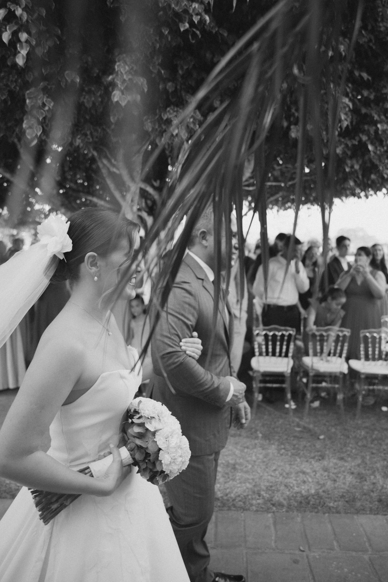 A black and white wedding ceremony photo with sun flare and guests in the background.