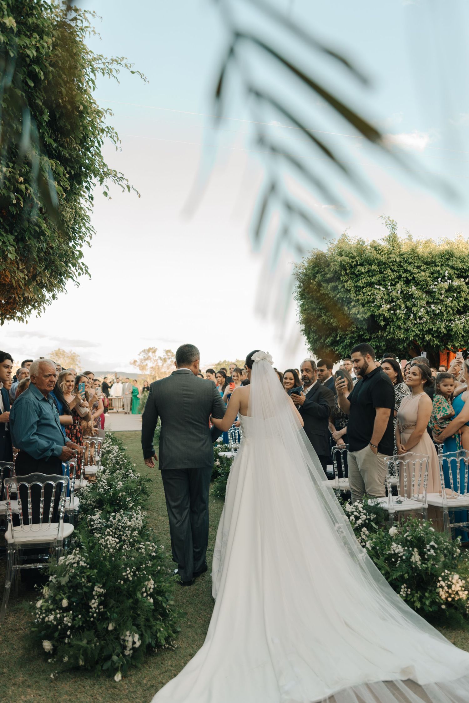 A long trailing wedding dress train as couple walks down an outdoor aisle lined with flowers.