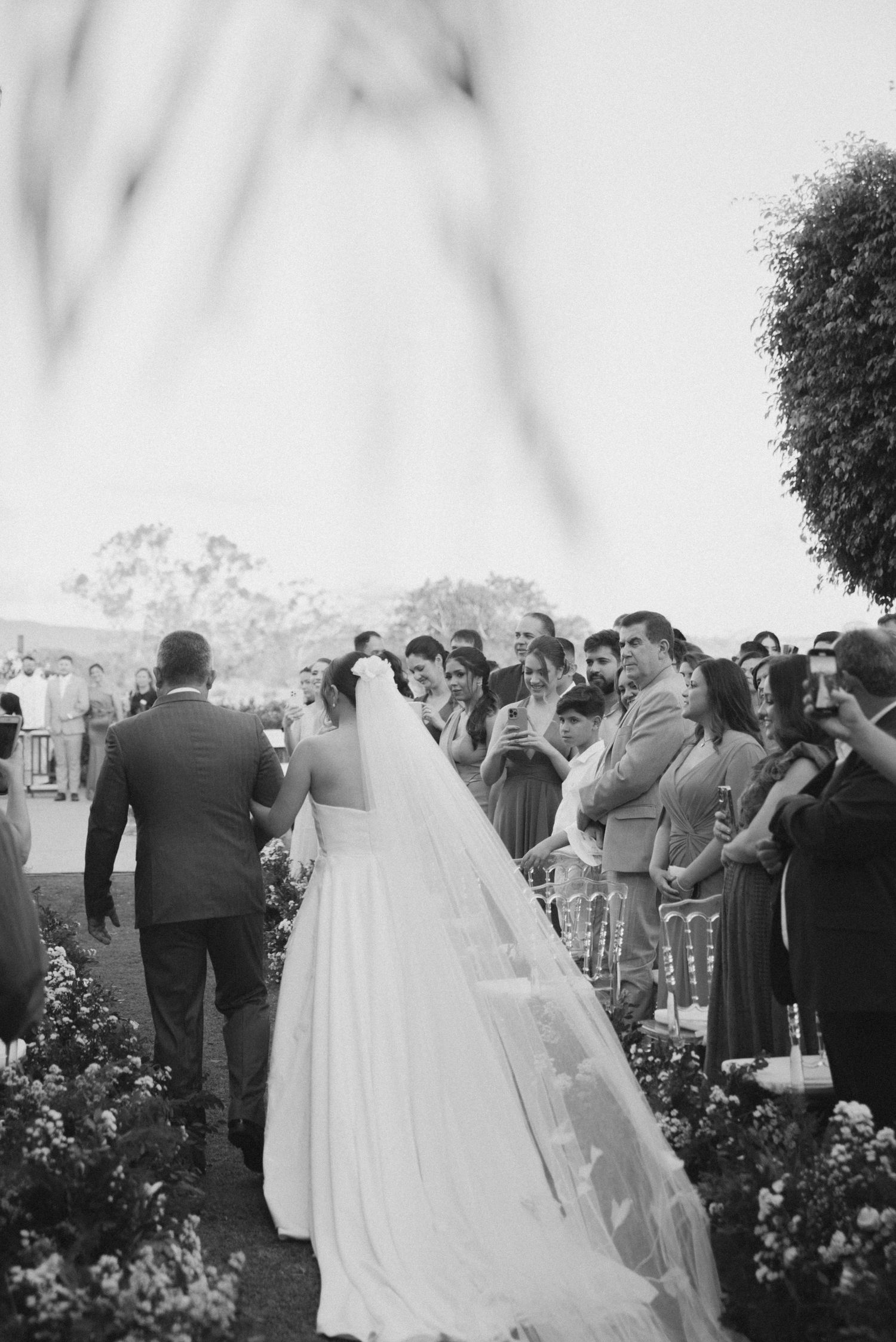 A black and white photo of a wedding ceremony with guests watching as a couple walks down a flower-lined aisle.