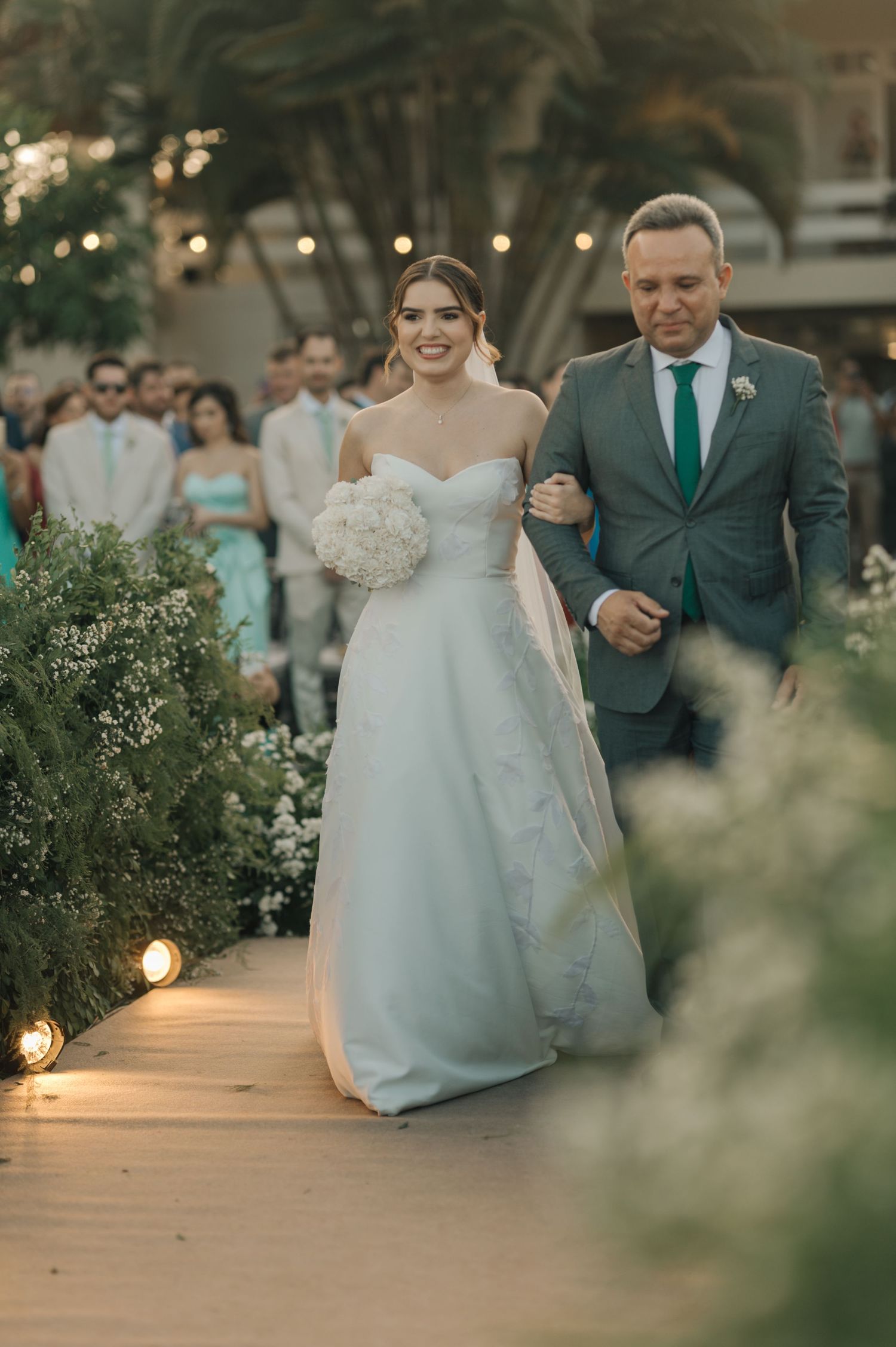 A wedding procession walking down an illuminated outdoor pathway lined with flowers at dusk.