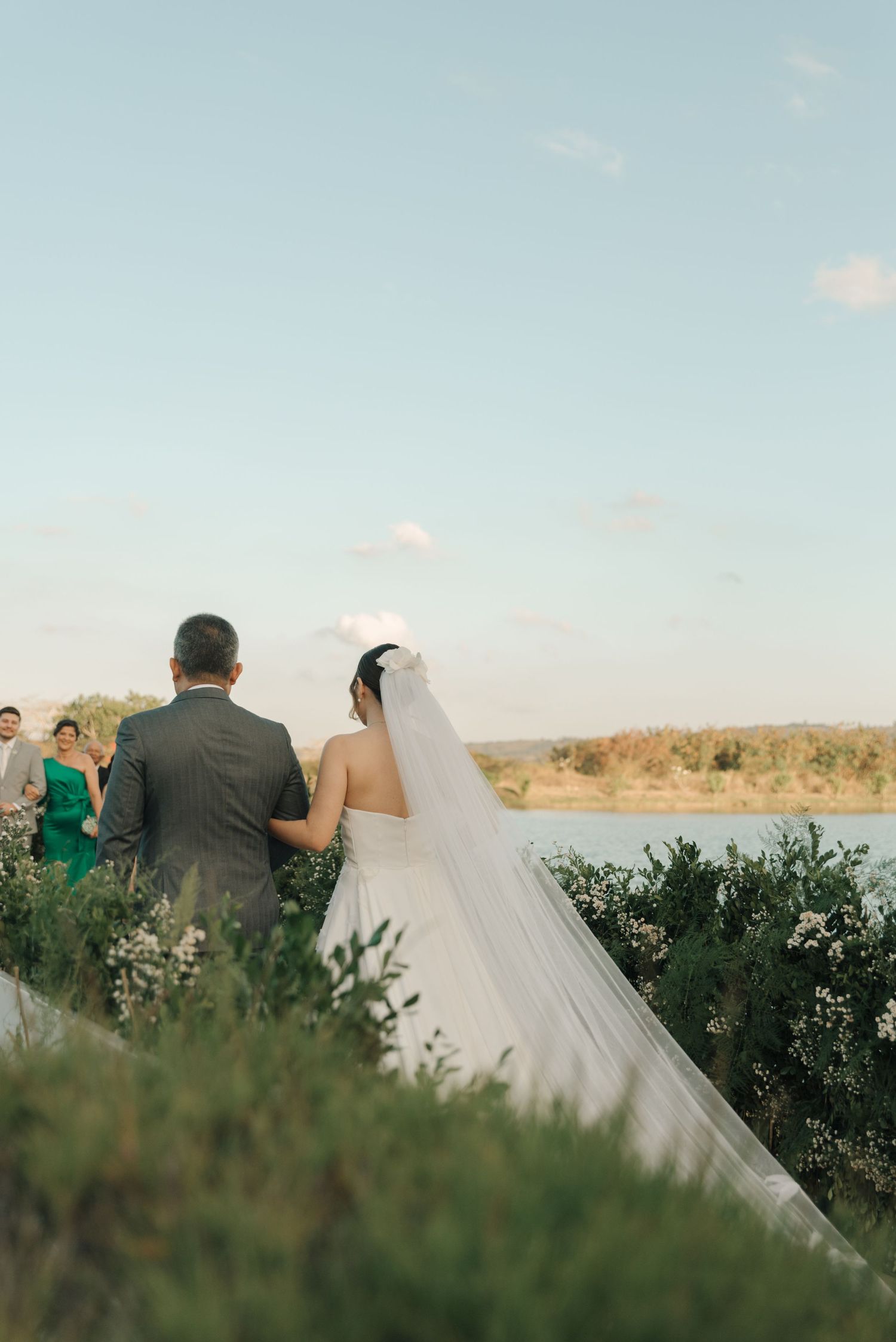 A couple in wedding attire walking along a scenic overlook with water and greenery in the background.