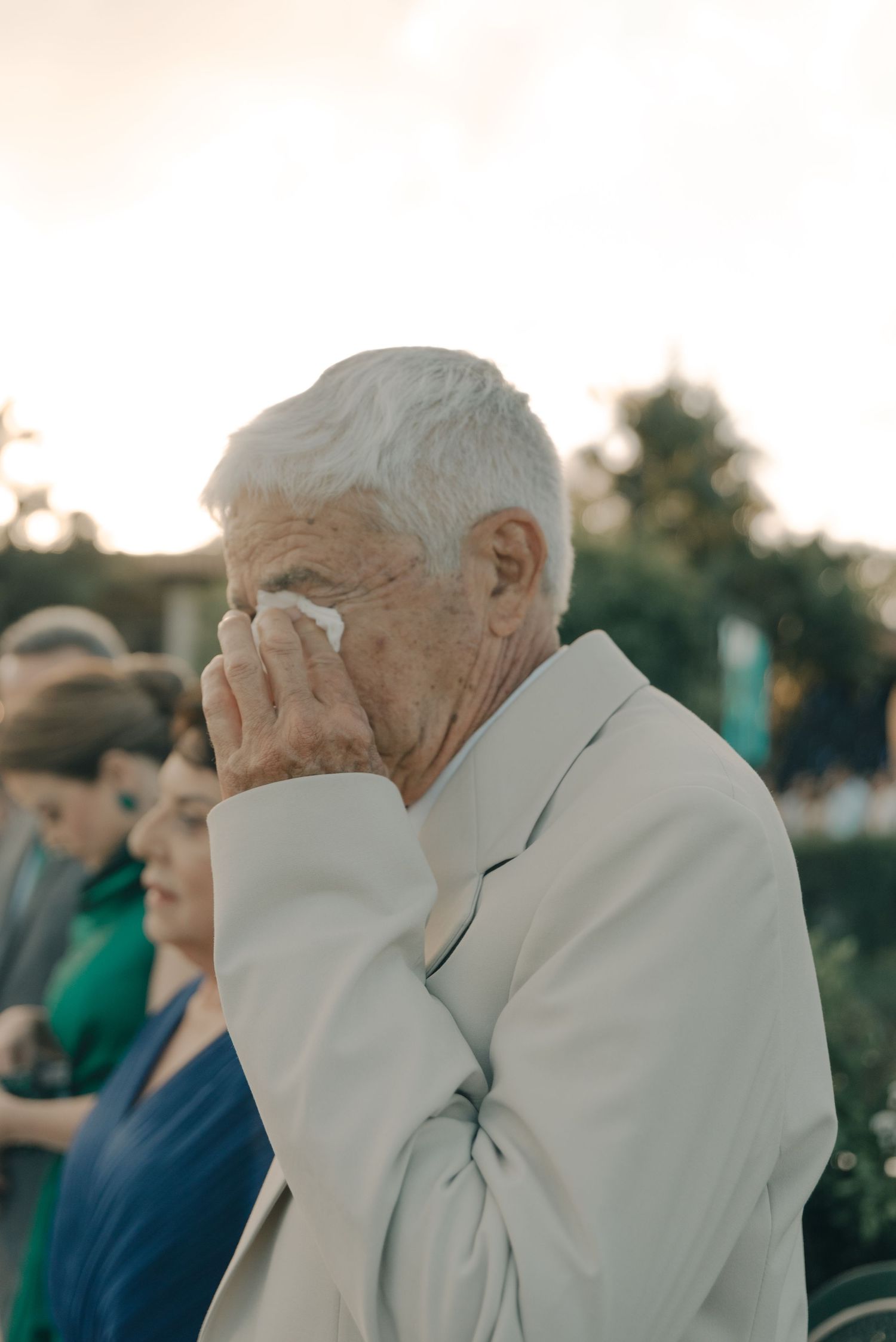 An elderly person in a white jacket wipes their eyes during an emotional outdoor ceremony.