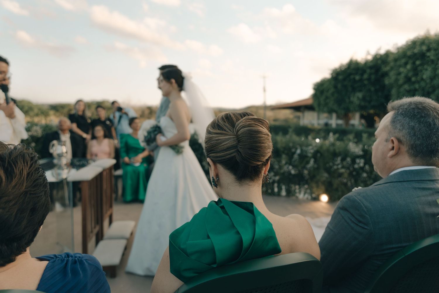 Wedding guests watch as a ceremony takes place outdoors at sunset.