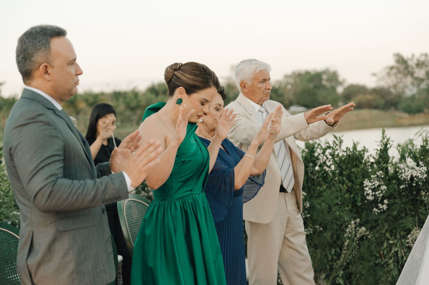 Wedding attendees in formal attire clap during an outdoor celebration near a waterfront setting.