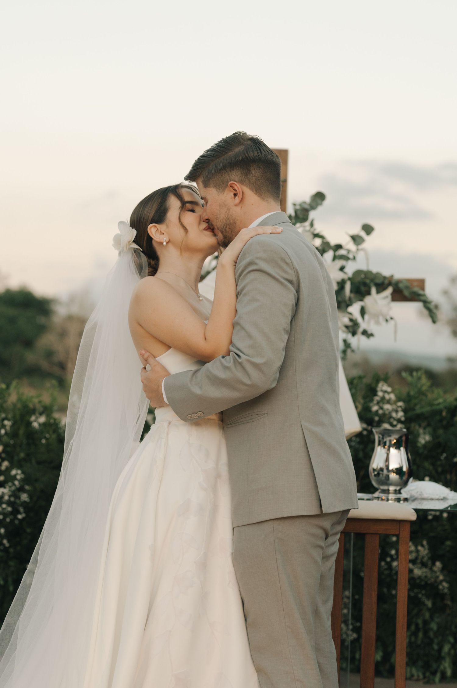 Bride in white gown and groom in grey suit embrace during their outdoor wedding ceremony.