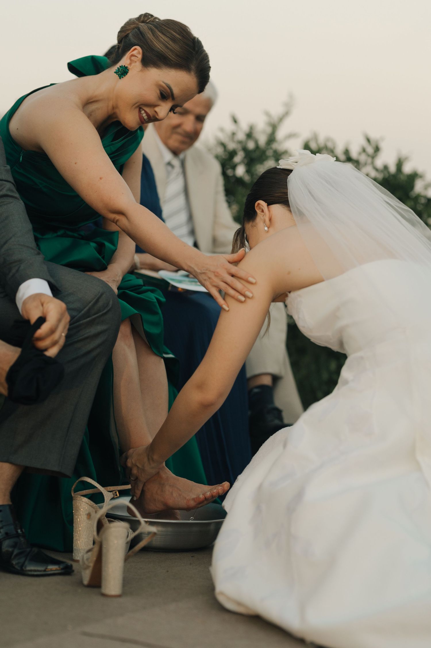 Bridesmaid in emerald dress helps bride with her shoe during wedding celebration.