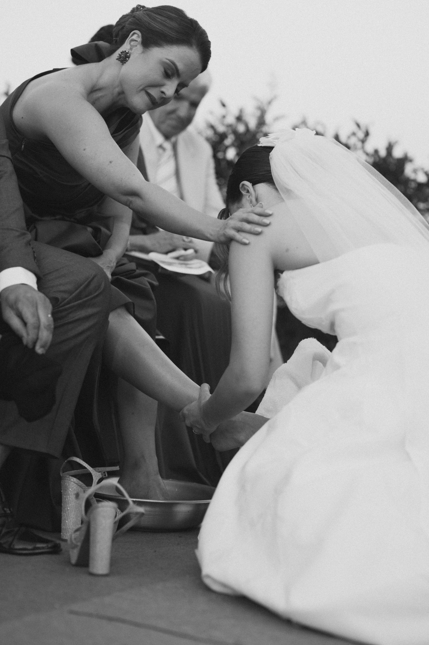 Black and white image of bridesmaid helping bride with her shoe during wedding celebration.