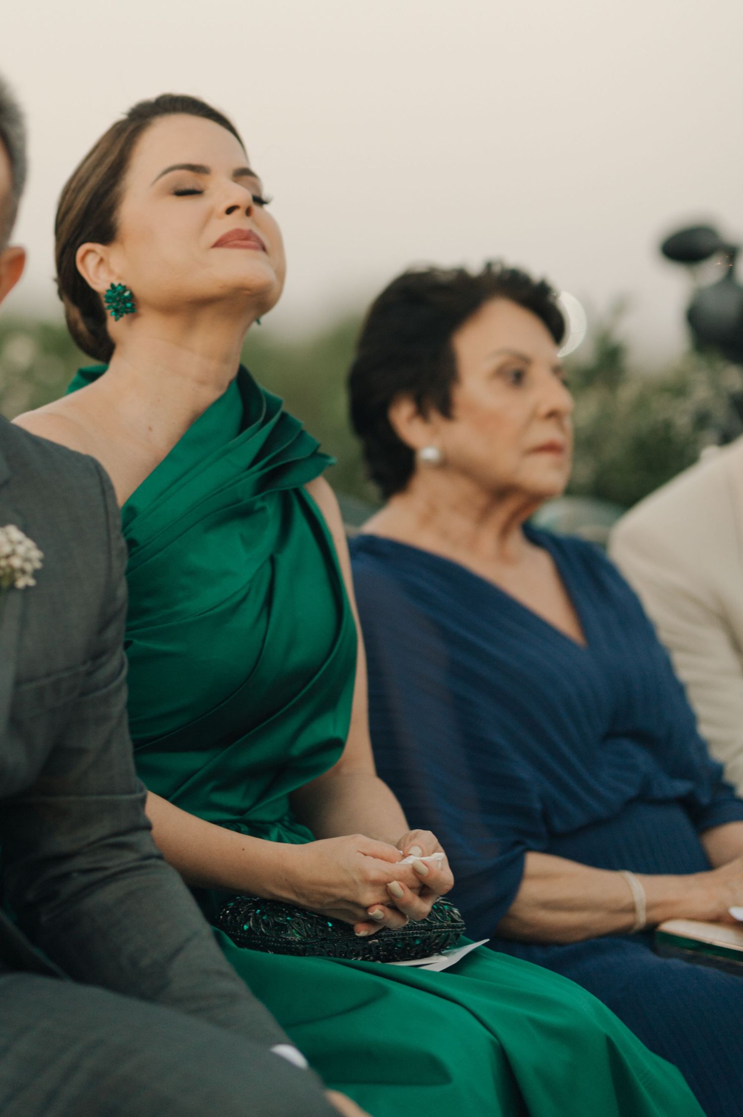 Wedding guests in elegant dresses sit attentively during outdoor ceremony.